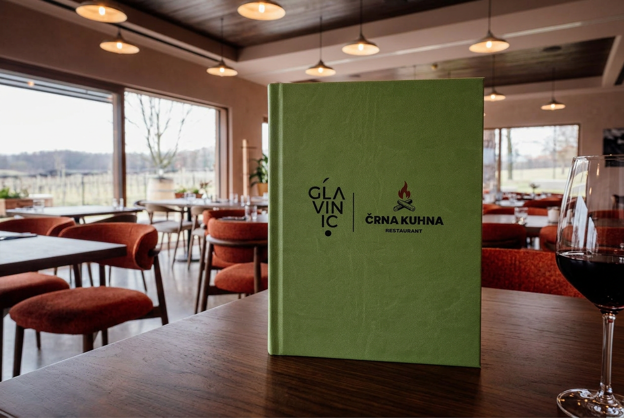 Zelene platnice menija Glavinič Črna Kuhna restaurant stojijo na temni leseni mizi v restavraciji Črna Kuhna, ob kozarcu rdečega vina, v ozadju mehko zamegljena notranjost z rjastimi bouclé stoli.