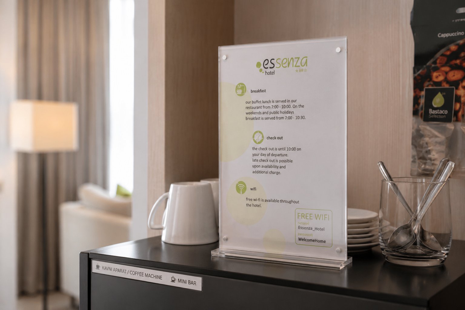 Akrilno namizno stojalo z informacijami hotela Essenza o zajtrku, odjavi in Wi-Fi povezavi, postavljeno na črni omarici poleg skodelic in pribora v hotelski sobi.