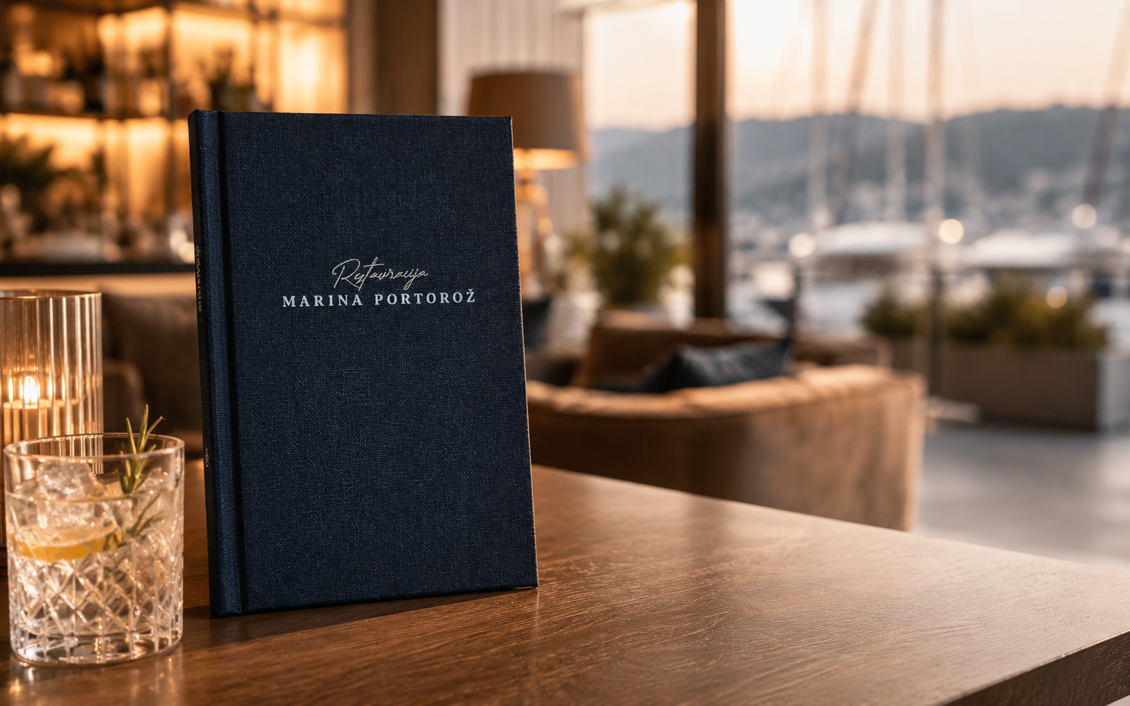 Temno moder premium meni Marina Portorož z elegantno srebrno gravuro stoji na leseni mizi v luksuznem hotelskem lounge baru ob marini, ob cocktail kozarcu in nežni sveči v toplem golden hour ambientu.