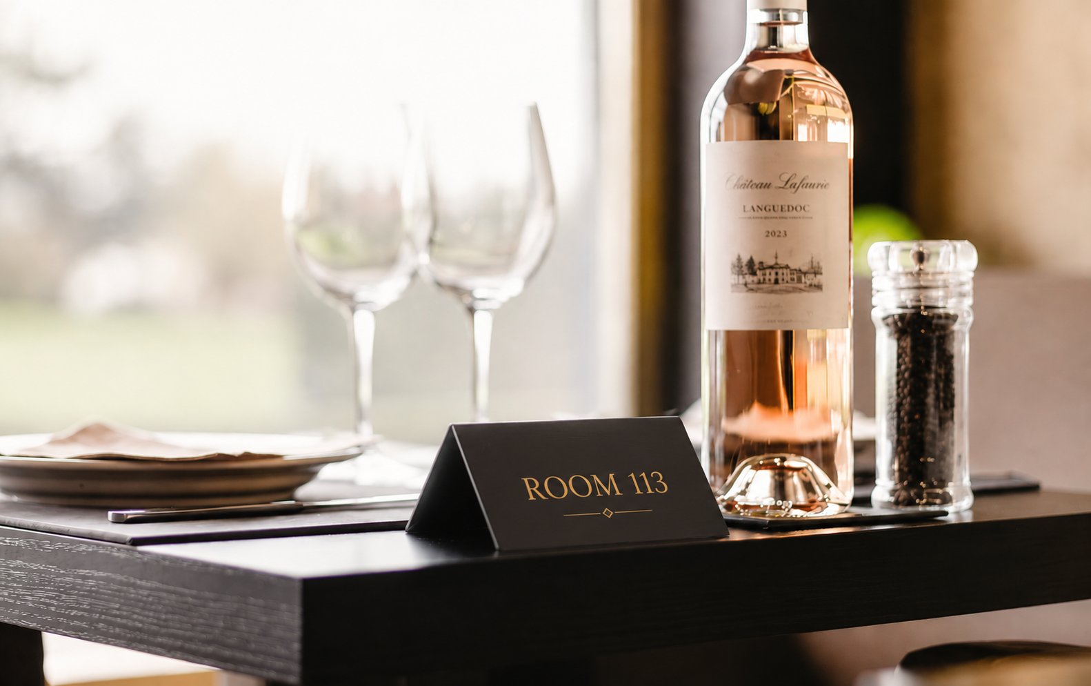 Črna kovinska rezervacijska tablica z zlatim napisom ROOM 113 na črni leseni mizi, ob steklenici roze vina Château Lafaurie, dveh kozarcih za vino in mlinčku za poper v elegantnem ambientu restavracije